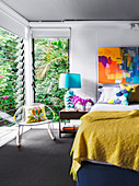 Double bed with mustard yellow bedspread, bedside table with blue table lamp and rocking chair in the bedroom, colorful artwork on the wall