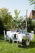 Gartentisch mit weißer Tischdecke, blauen Akzenten und Lichterketten im Sommergarten