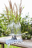 Glasvasen mit Ziergräsern und Artischocken auf Holztisch im Garten