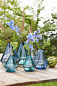 Blaue Glasvasen mit Rittersporn  und Artischocken auf Holztisch im Garten