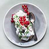 A slice of Pavlova, topped with redcurrants and raspberries, decorated with thyme