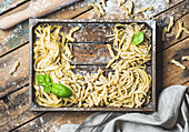 Various homemade fresh uncooked Italian pasta with flour and green basil leaves in wooden tray