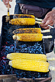 A Balinese street vendor selling spicy corn cobs on a charcoal bbq with a cigarette in the hand