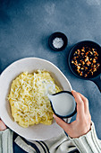 Hands pouring milk from a black ceramic cup on mashed potatoes and grated cheese in a white ceramic bowl