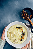 Hands holding cheesy mashed potatoes with roasted walnuts and black pepper in a white ceramic bowl