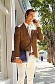 A young businessman wearing a brown blazer and light trousers walking down the road on the phone