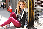 A young blonde woman wearing a jacket and dark red trousers sitting on a pavement