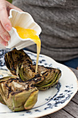 A woman pouring melted garlic butter onto grilled artichokes on a blue and white platter