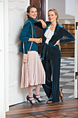 Two women wearing petrol autumnal outfits: a jumper with a pleated skirt, and a long velvet blazer with a ruffled blouse and velvet trousers