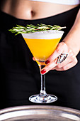 A cocktail with an egg white topping and rosemary being served