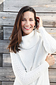 A brunette woman wearing a white knitted jumper standing in front of a wooden wall