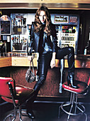 A young woman sitting on a bar wearing black leather and a jacket
