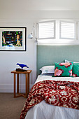 Bed with a high mint green headboard under a narrow, open louvre window