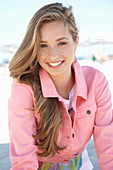 A young blonde woman on a beach wearing a t-shirt and a pink denim jacket