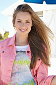A young blonde woman on a beach wearing a printed t-shirt and a pink denim jacket