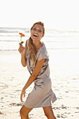 A mature blonde woman on a beach wearing a silver summer dress and holding an orange flower