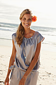 A mature blonde woman on a beach wearing a silver summer dress with an orange flower behind her ear