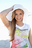 A young blonde woman on a beach wearing a colourful t-shirt and a white summer hat