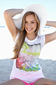 A young blonde woman on a beach wearing a colourful t-shirt, a white summer hat and pink shorts
