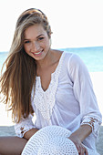 A young blonde woman on a beach wearing a white summer dress and a white summer hat