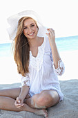 A young blonde woman on a beach wearing a white summer dress and a white summer hat