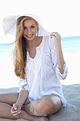 A young blonde woman on a beach wearing a white summer dress and a white summer hat