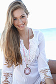 A young blonde woman on a beach wearing a white summer dress and a pink necklace