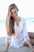 A young blonde woman on a beach wearing a white summer dress and a pink necklace