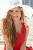 A young blonde woman on a beach wearing a red summer dress and a white hat