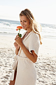 Reife blonde Frau in Dessous und Strickjacke und mit Blume in der Hand am Strand