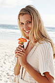 A mature blonde woman on a beach wearing lingerie and a cardigan and holding a flower