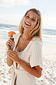 A mature blonde woman on a beach wearing lingerie and a cardigan and holding a flower