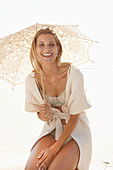 A mature blonde woman on a beach wearing lingerie and a cardigan with a parasol