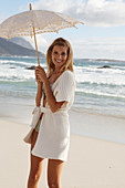 A mature blonde woman on a beach wearing lingerie and a cardigan with a parasol