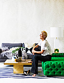 Blond woman sitting on green ottoman, gold-colored coffee table and gray sofa in living room