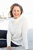 A brunette woman on a beach wearing a white knitted jumper and dark trousers