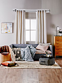Graues Polstersofa mit Decken und Kissen vor Fenster, Sitzkissen und Stuhl als Aufbewahrungsmöbel auf Teppich