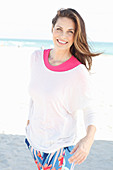 Brünette Frau in pinkfarbenem T-Shirt, weißem Pulli und bunter Hose am Strand