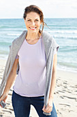 Brunette woman wearing lilac T-shirt, jeans and grey sweater over shoulders on beach