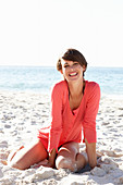 A mature brunette woman on a beach wearing a black bikini and a red shirt