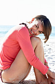 A mature brunette woman on a beach wearing a black bikini and a red shirt