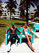 A young blonde woman wearing a bathing suit and a man wearing shorts sitting on chairs