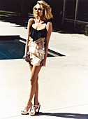 A young blonde woman posing in the garden of a house with a pool wearing a black-and-beige evening dress