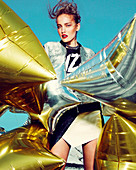 Junge Frau in silberner Jacke und schwarz-weißem Rock mit goldenen und silbernen Luftballons