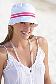 A young brunette woman on a beach wearing a white top and a white hat