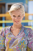 A mature blonde woman with short hair on a beach wearing a patterned summer dress