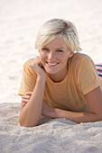 A mature blonde woman with short hair on a beach wearing a striped bikini and an orange top