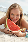 A young blonde woman on a beach wearing a colourful summer dress holding a wedge of melon