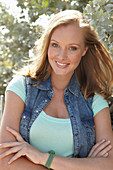 A young blonde woman outside wearing a sleeveless denim jacket and a light-blue top