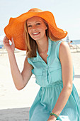 A young blonde woman on a beach wearing a light-blue summer dress and an orange summer hat